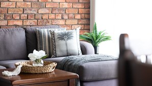 Un salon avec un canapé gris, des coussins à motifs, un panier en osier contenant une fleur blanche et un mur en briques.