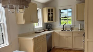 A spacious kitchen with wooden flooring, white cabinetry, a large window, and a hanging light fixture.