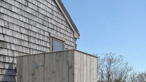 A wooden outhouse with a simple design, situated on a grassy area in front of a house with a sloped roof.