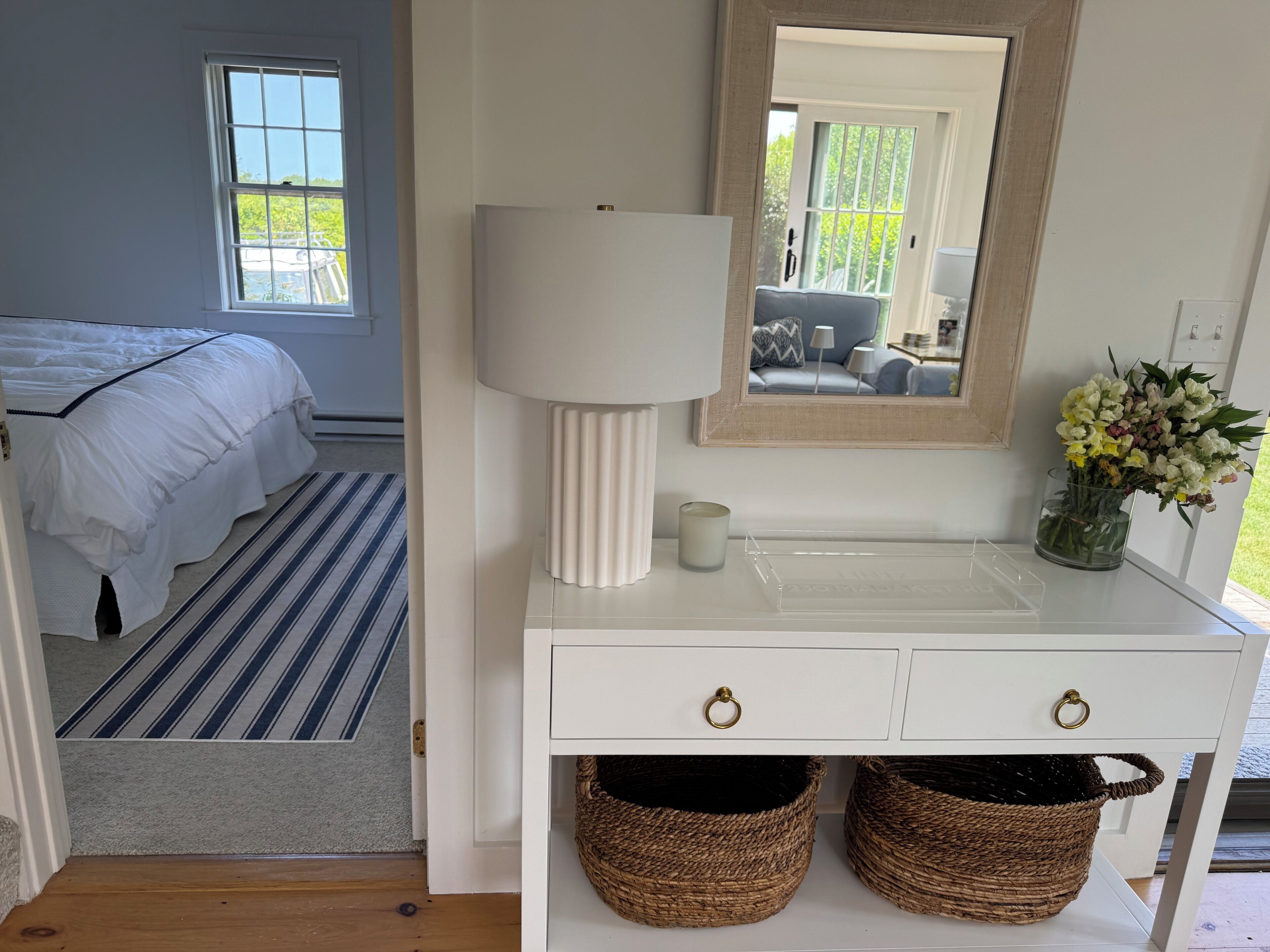 Ein Schlafzimmer mit einem Bett, einem Nachttisch mit Lampe, einem Spiegel und einer Vase mit Blumen.