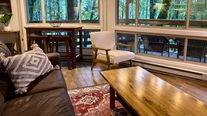 Een gezellige woonkamer met een houten salontafel, een bank en een fauteuil. Grote ramen bieden uitzicht op een bebost gebied.