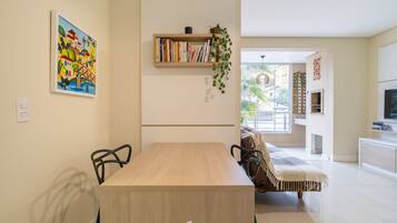 Un comedor moderno con una mesa y sillas de madera, una estantería con libros y una planta colgante, y un cuadro colorido en la pared.