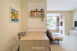A modern dining area with a wooden table and chairs, a bookshelf with books and a hanging plant, and a colorful painting on the wall. - Apart With Barbecue 600m From the Beach Rpn0101 (Florianópolis)