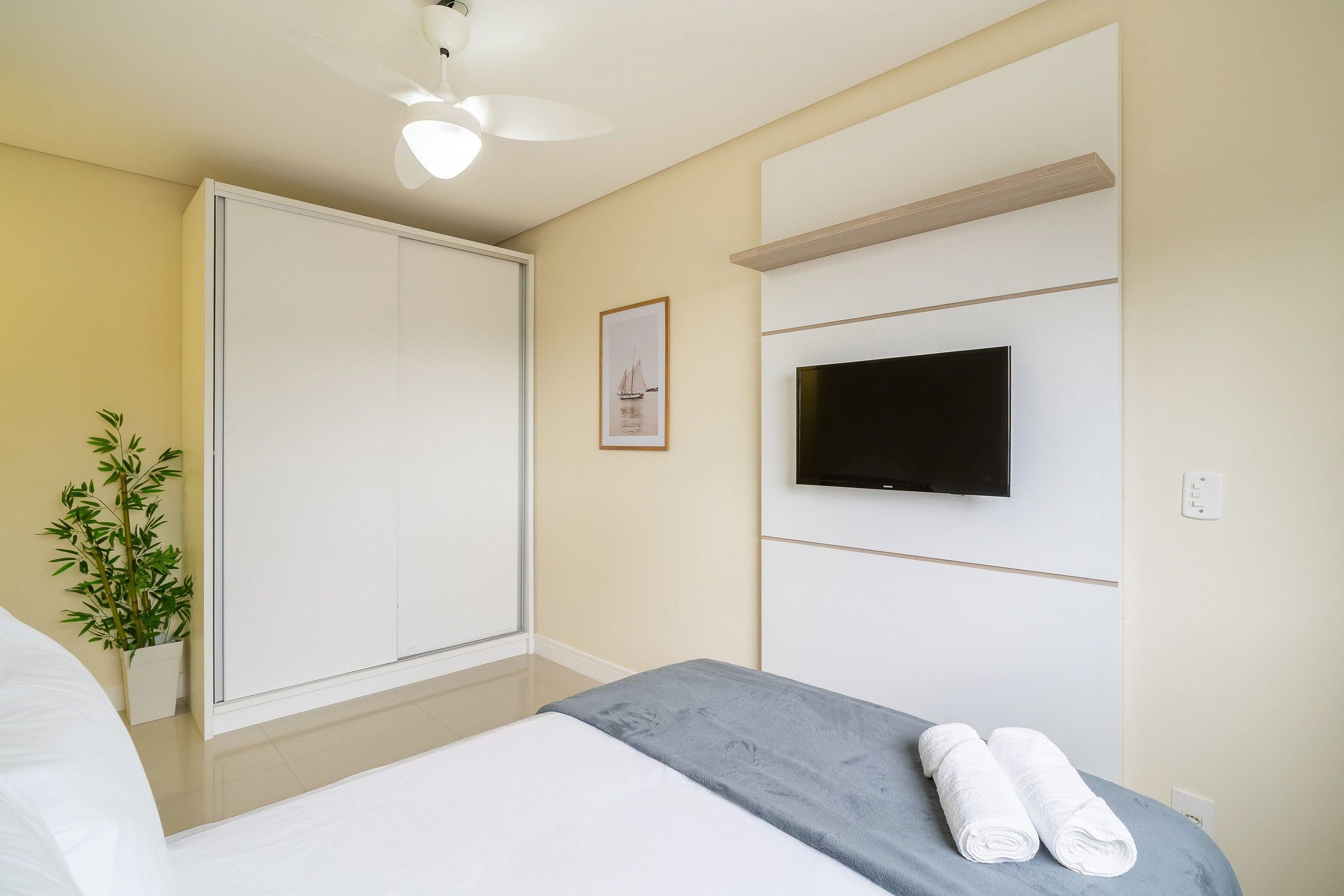 A hotel room with a bed, a TV mounted on the wall, a wardrobe, and a potted plant.