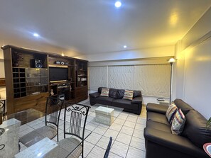 A modern living room with a glass coffee table, a black leather sofa, and a wooden entertainment unit with shelves and a TV. - Aparts4Days 1-Bedroom Loft in Downtown Miraflores Near the Malecón (Lima)