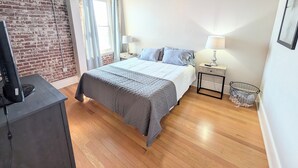 A bedroom with a bed, a desk, a TV, a ceiling fan, a window with curtains, and a brick wall.