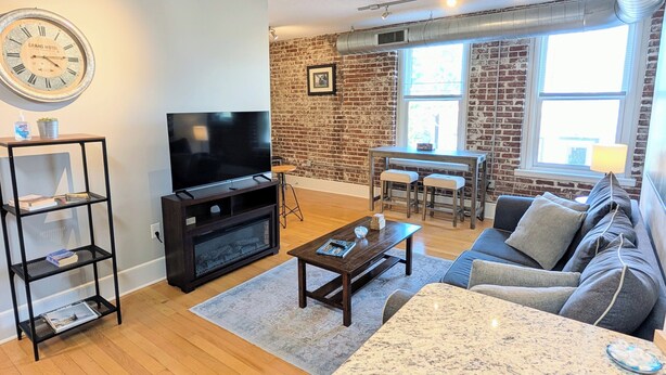 A living room with a brick wall, a television, a coffee table, a sofa, and a dining area with two stools.