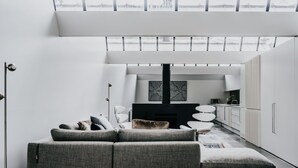 Un salon moderne avec un canapé gris, un meuble de rangement noir pour appareils électroniques et un coin cuisine doté de meubles blancs et d’un four à micro-ondes.