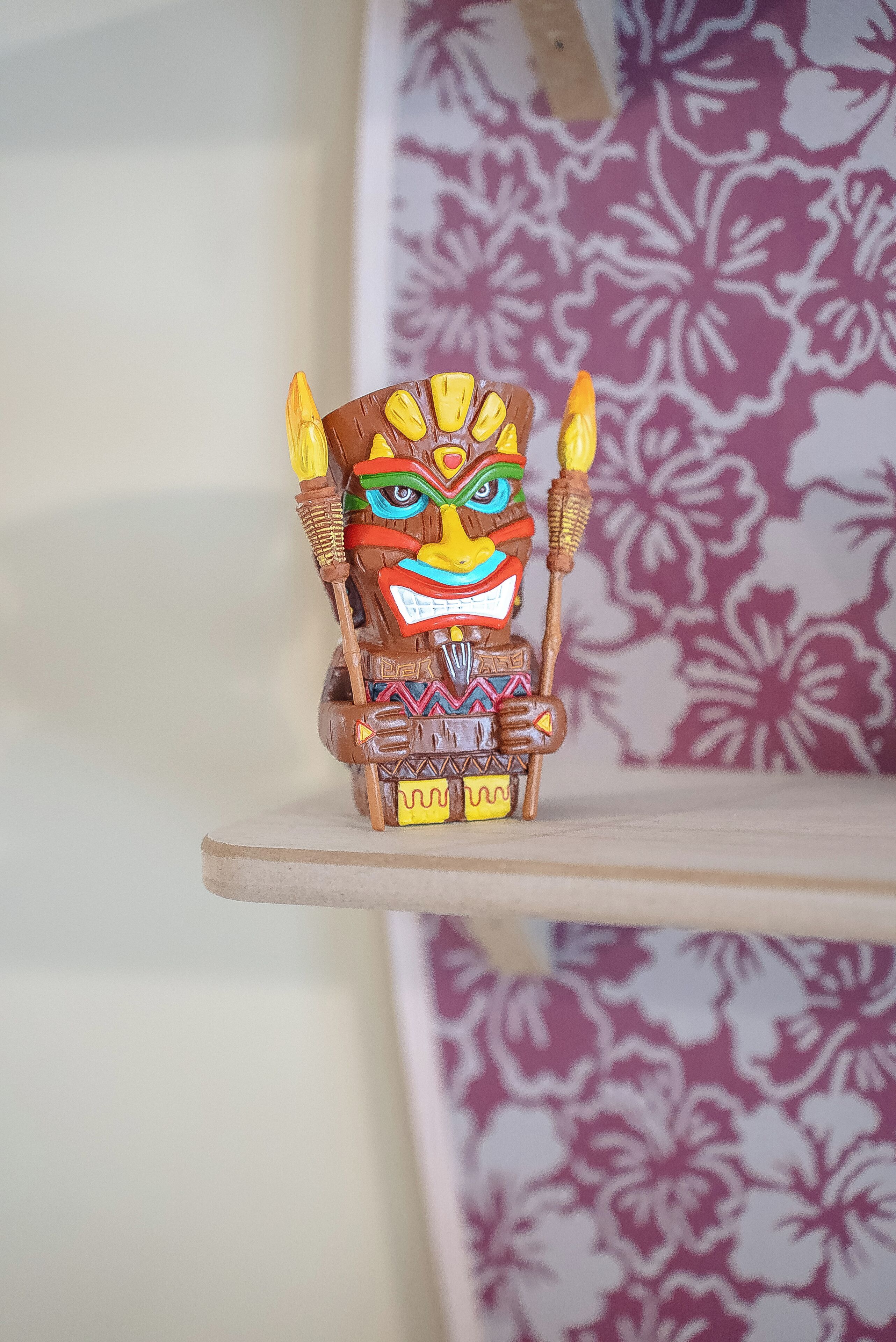 Eine dekorative Holzfigur mit farbenfrohem Gesicht und Stab, platziert auf einem Regal vor einer Wand mit Muster.