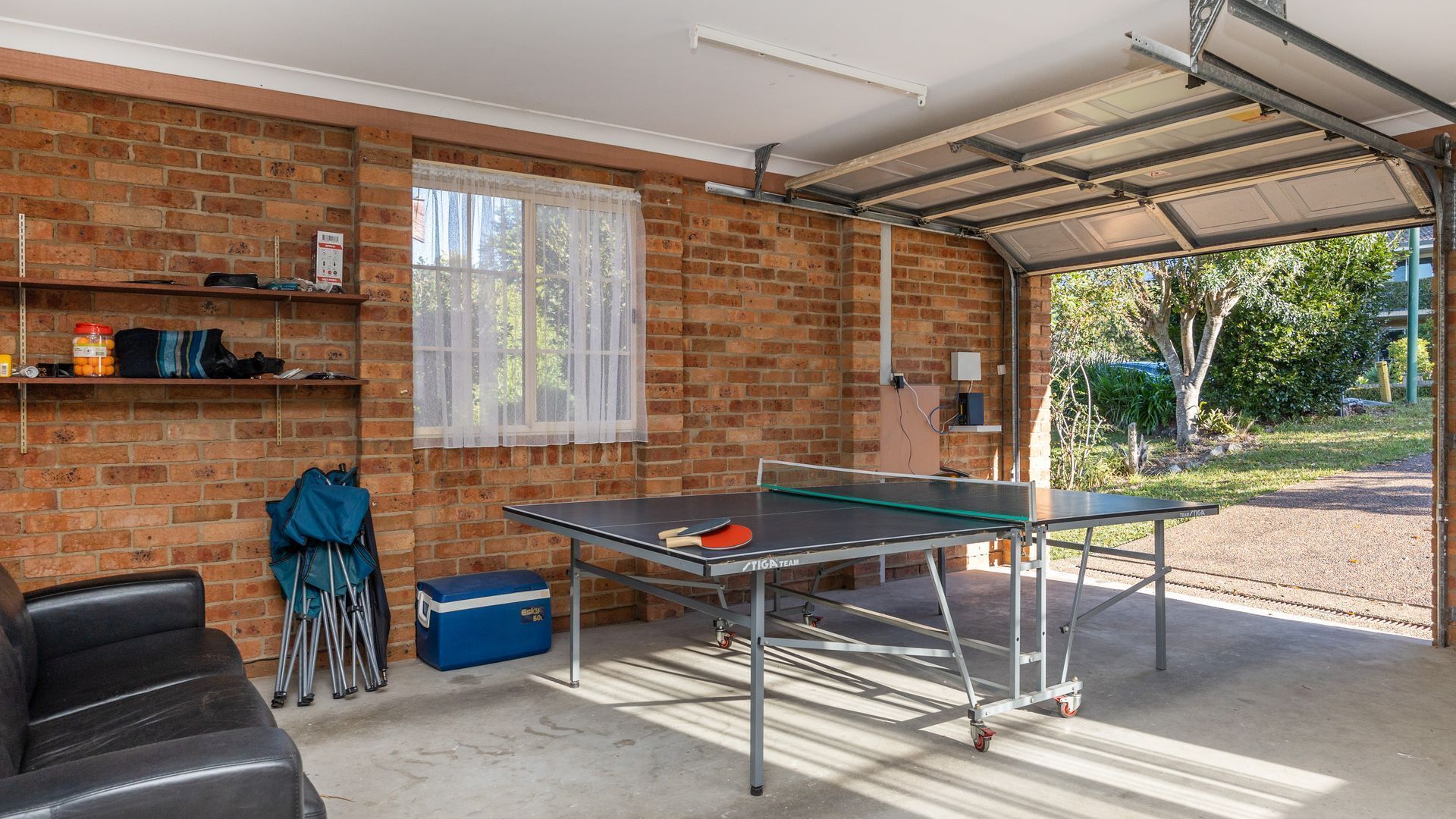 A ping pong table in a garage with a brick wall, a window with sheer curtains, and a blue cooler on the floor.