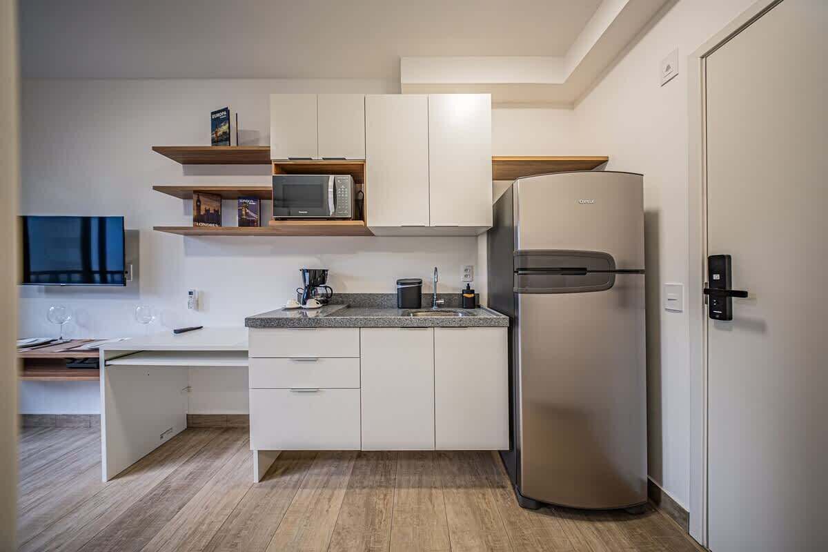 Une cuisine compacte avec des meubles blancs, un four à micro-ondes encastré, un réfrigérateur en acier inoxydable et un plan de travail sur lequel se trouve une machine à café.