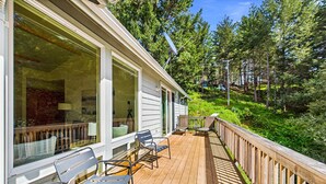A wooden deck with outdoor seating, large windows, and a view of a forested hillside. - Coastal Canopy-Secluded home with ocean view and jetted tub. Sleeps 6 (Whitethorn)