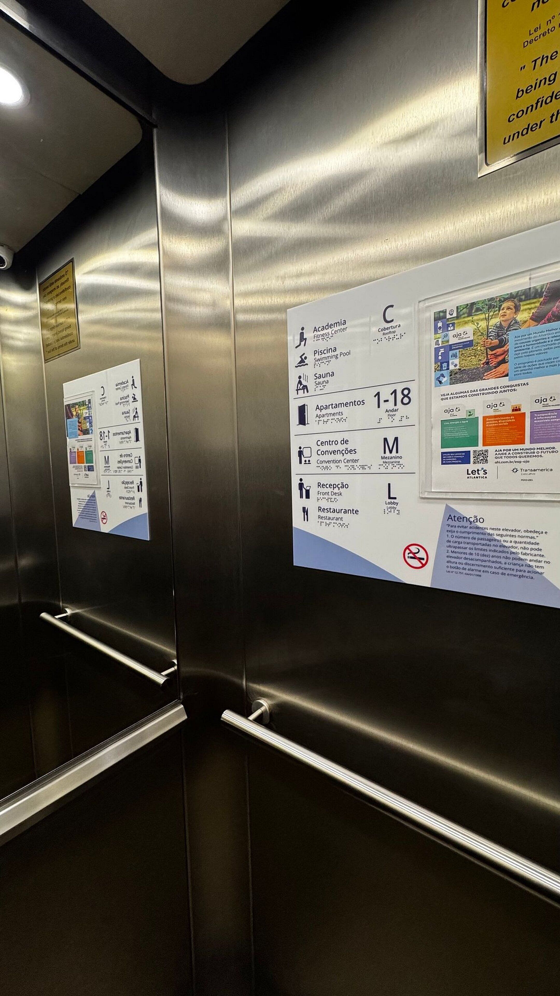 Interior de un ascensor con paredes y pasamanos metálicos. Hay carteles informativos en las paredes que indican servicios como gimnasio, piscina, sauna, apartamentos, centro de convenciones, vestíbulo y restaurante.