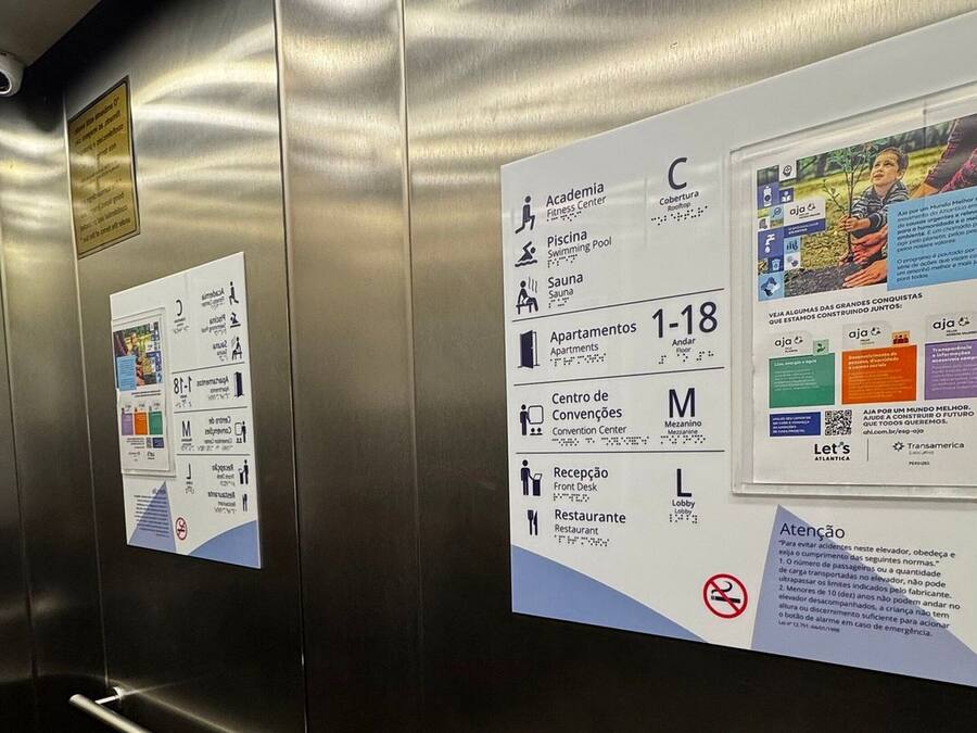 Interior de un ascensor con paredes y pasamanos metálicos. Hay carteles informativos en las paredes que indican servicios como gimnasio, piscina, sauna, apartamentos, centro de convenciones, vestíbulo y restaurante.