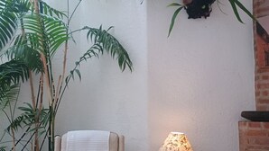 A cozy living room with a beige armchair, a glass coffee table with magazines, a lamp, and large potted plants.