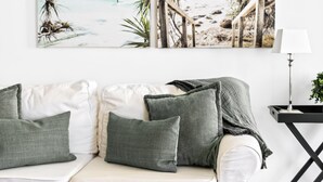 Un canapé blanc avec des coussins verts, un tableau sur le thème de la plage au-dessus, et une lampe sur une table d’appoint.