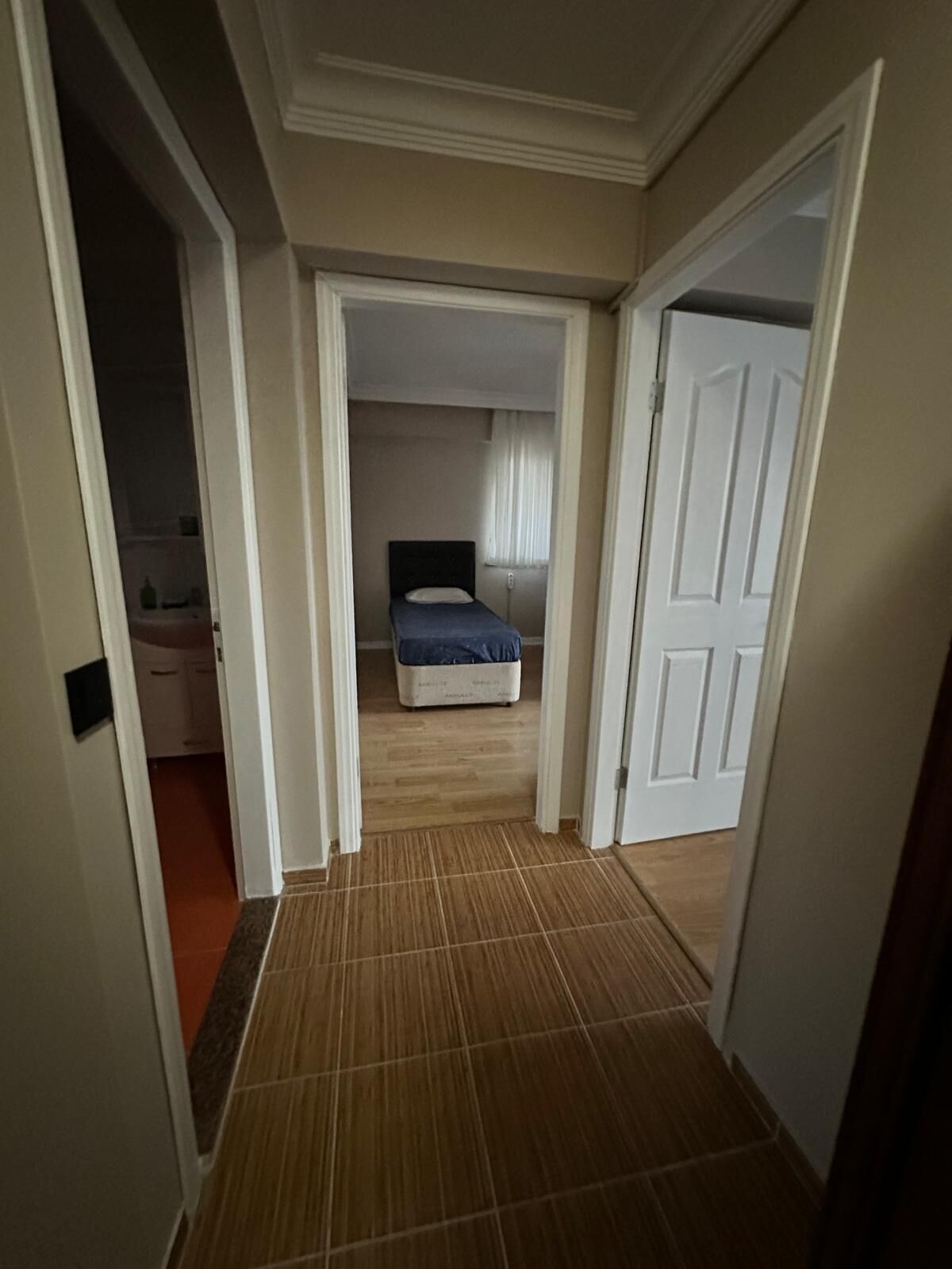 A narrow hallway with a wooden floor leading to a bedroom with a bed and a nightstand.