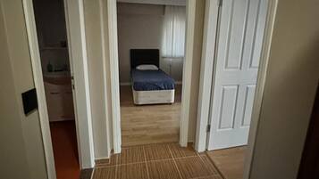 A narrow hallway with a wooden floor leading to a bedroom with a bed and a nightstand.