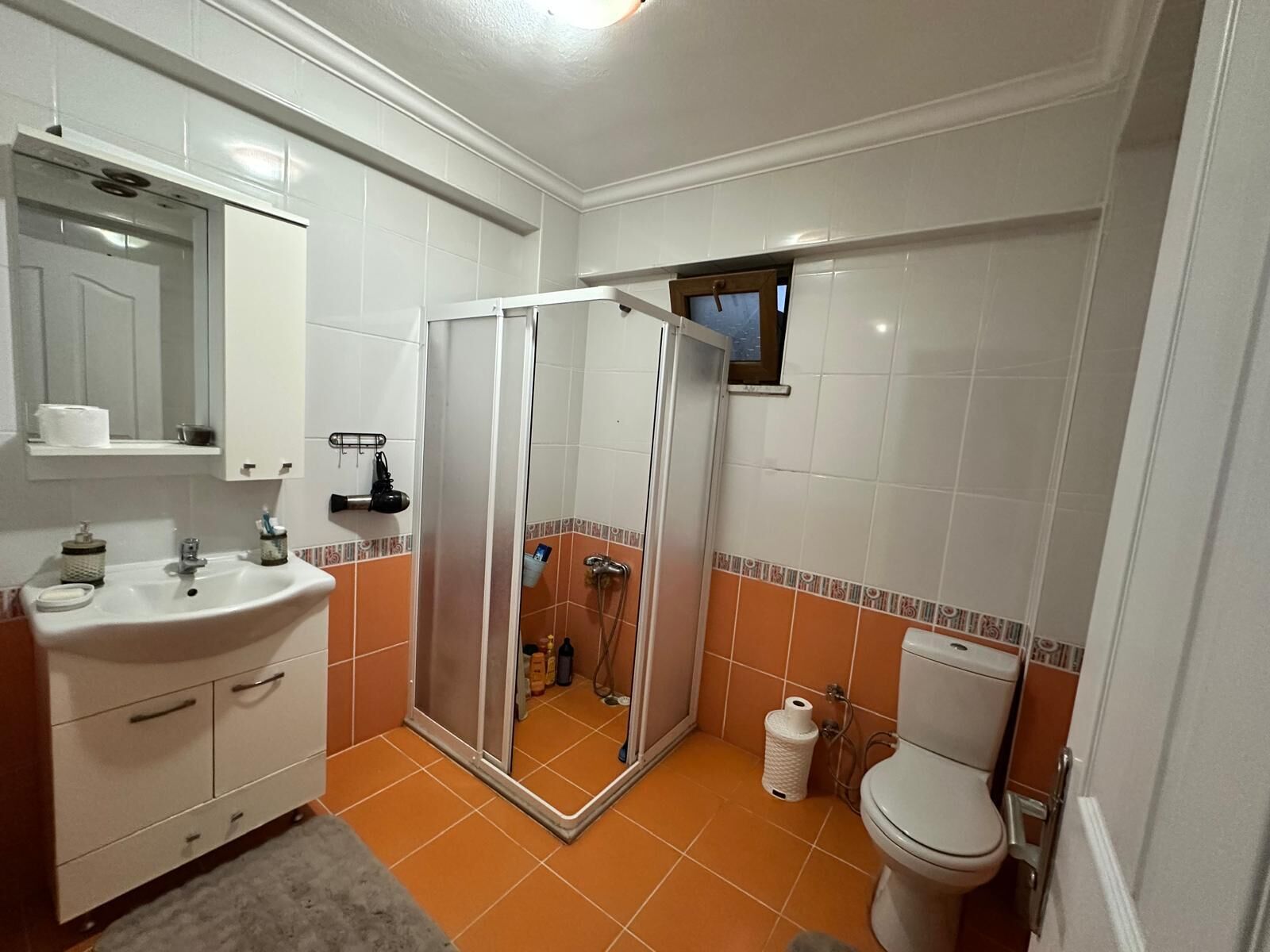Un baño con lavamanos, espejo y inodoro blancos, cabina de ducha y paredes con azulejos anaranjados.