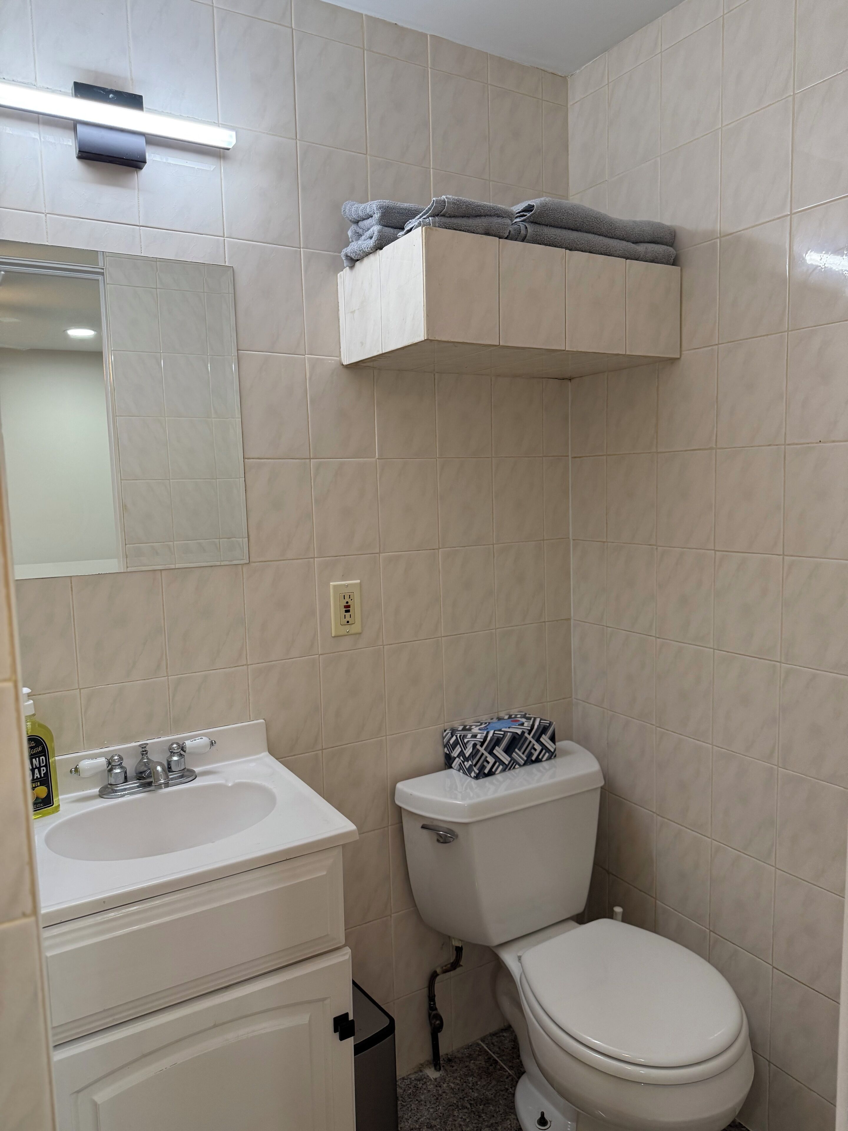 A bathroom with a white sink, mirror, and toilet. There is a towel rack above the toilet and a bottle of hand soap on the sink.