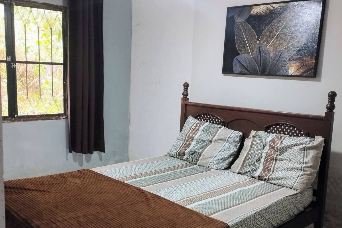 A bedroom with a bed, a ceiling fan, a window with curtains, and a wall-mounted painting.