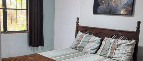 Une chambre à coucher avec un lit, un ventilateur de plafond, une fenêtre avec des rideaux et un tableau accroché au mur.