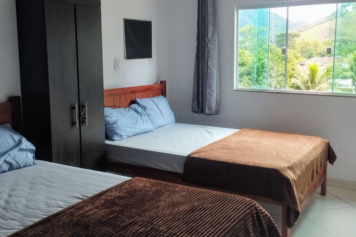 A room with two beds, a wardrobe, a ceiling fan, a window with a view of greenery, and a curtain.