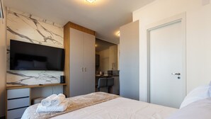 Une chambre d’hôtel moderne avec un mur décoré d’un motif imitant le marbre, un lit avec des serviettes soigneusement pliées, un bureau doté d’un téléviseur à écran plat et un coin repas séparé, visible à travers une porte ouverte.