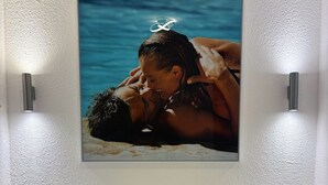 A bedroom with a bed, white pillows, a wall-mounted picture of a couple by the sea, and two wall sconces.