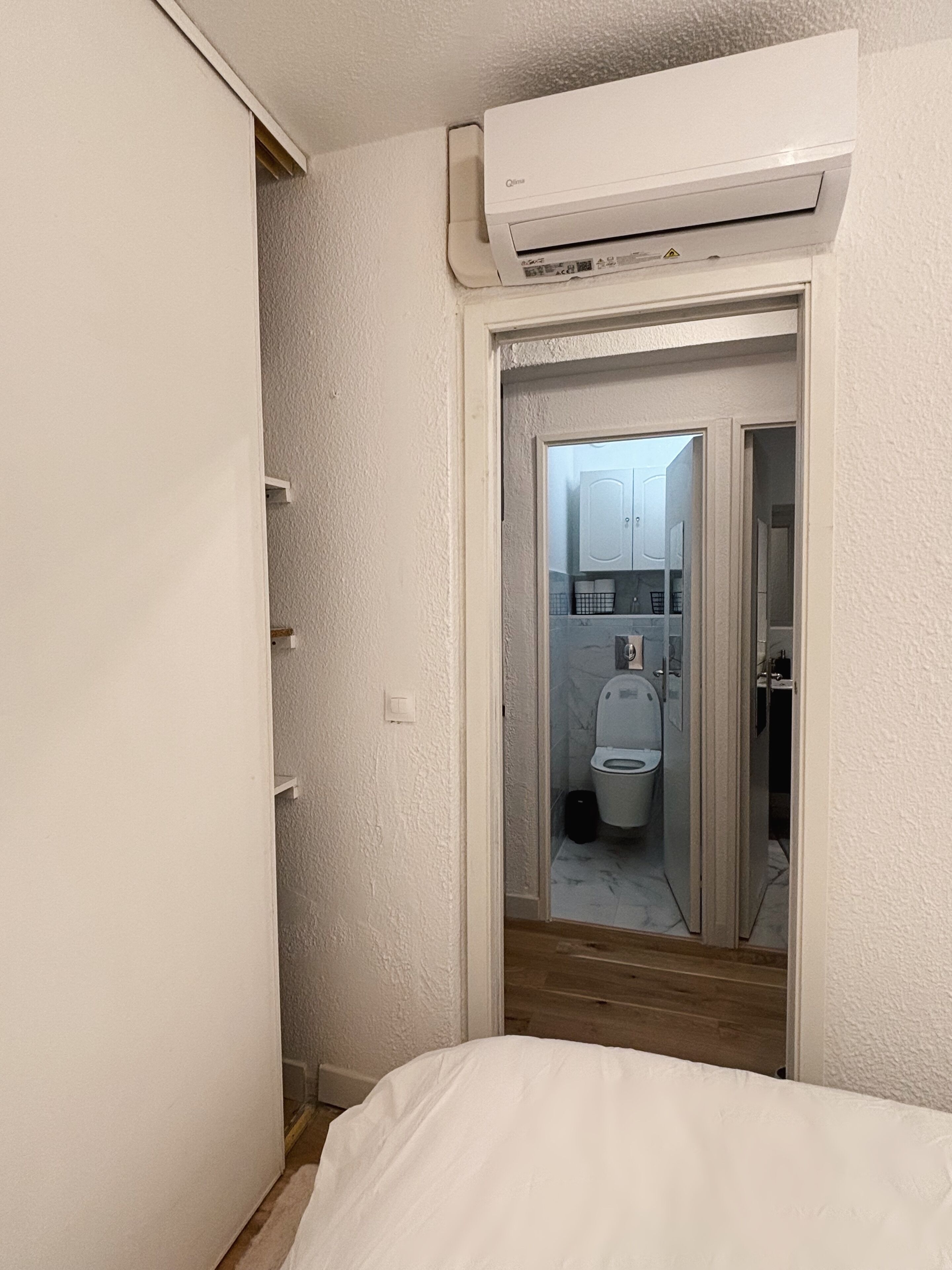 Una camera da letto con un letto, una parete bianca, una porta che conduce al bagno e un condizionatore d'aria posizionato sopra la porta.