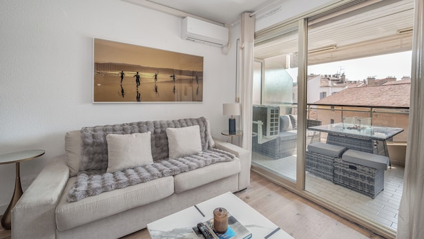 A modern living room with a sofa, a coffee table, and a large window overlooking a balcony with outdoor furniture.