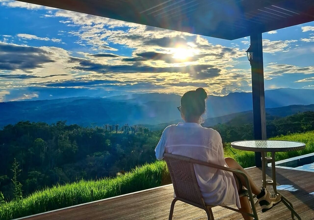 Iemand zit aan een tafel met uitzicht op een bergachtig landschap en de ondergaande zon.