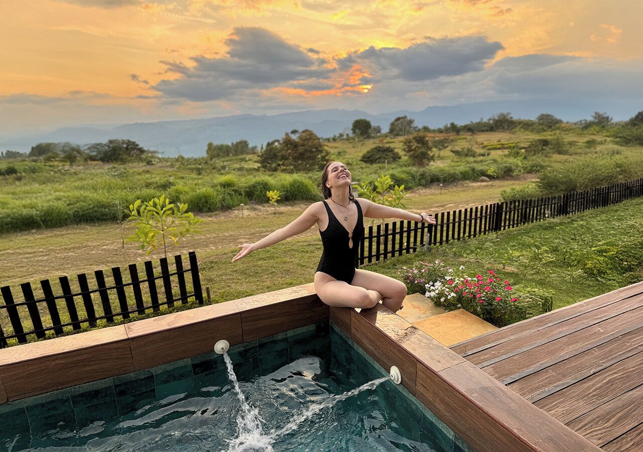 A person sitting on a wooden deck by a swimming pool with a sunset in the background.