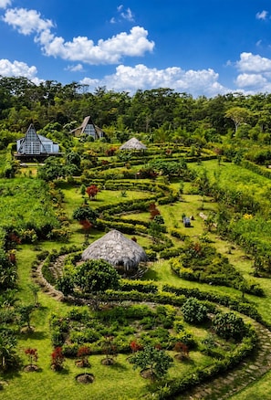 A lush garden with a thatched-roof structure, a blue building, and a white building with a triangular roof. - El Resort de Yanashpa - Tarapoto (Cacatachi)