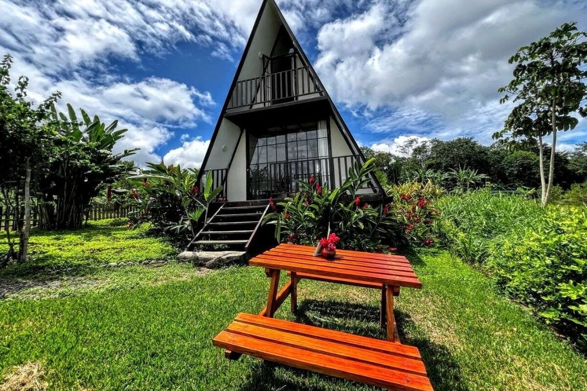 Un edificio triangular con balcón, rodeado de abundante vegetación y una mesa de picnic al frente.