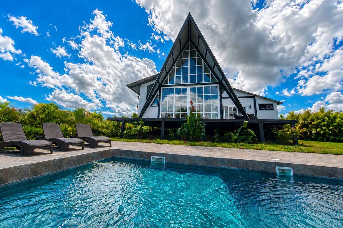Un edificio moderno con techo triangular, piscina y sillones de descanso.