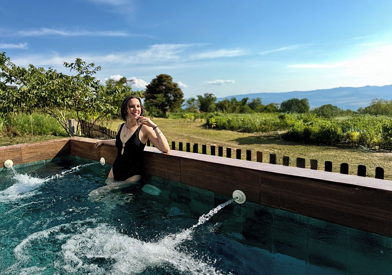 Una persona en traje de baño negro sentada en el borde de madera de una piscina con agua en movimiento, rodeada de vegetación y un cielo despejado.