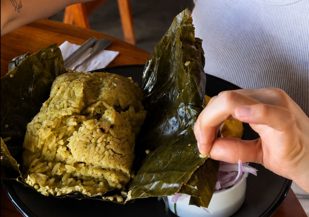 A person holding a leaf-wrapped food item, possibly a traditional dish, with a bowl of sauce or dip nearby.