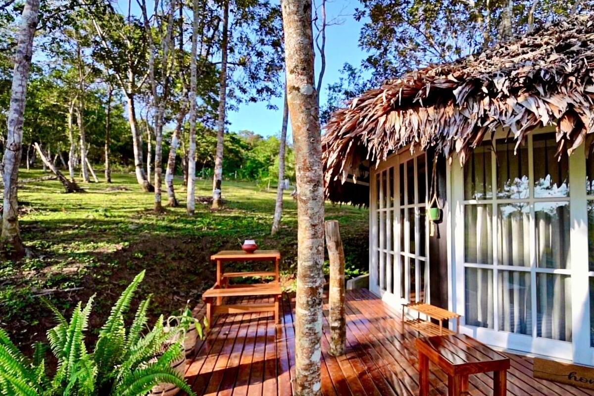 Una terraza de madera con una estructura de techo de paja, un banco y una mesa con un cuenco.