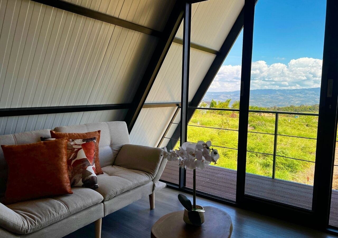Een moderne woonkamer met een bank, kussens en een bloemenvaas. Een groot raam biedt uitzicht op een groen landschap.