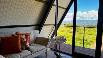 Een moderne woonkamer met een bank, kussens en een bloemenvaas. Een groot raam biedt uitzicht op een groen landschap.