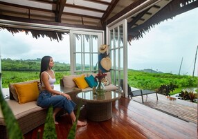 Una mujer sentada en un sofá con una mesa de vidrio en una habitación con ventanas amplias y vistas a un paisaje verde.