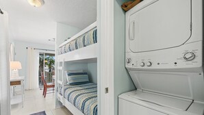 A laundry room with a stacked washer and dryer, a bunk bed with striped bedding, a small table with a lamp, and a view of palm trees through a window. - No Worries, Relaxing Sound side Retreat with pool (Navarre)