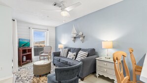 A living room with a grey sofa, a wooden chair, a small ottoman, a side table with a lamp, and a wall-mounted shelf with a sailboat decoration. - No Worries, Relaxing Sound side Retreat with pool (Navarre)