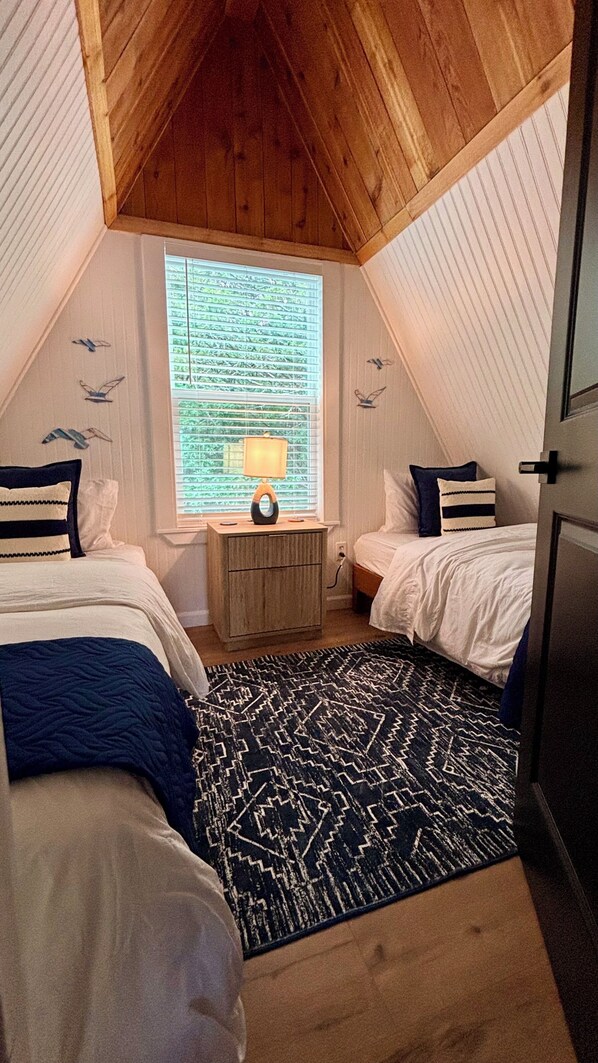 A cozy attic bedroom with two beds, a window with blinds, a lamp, and a patterned rug.