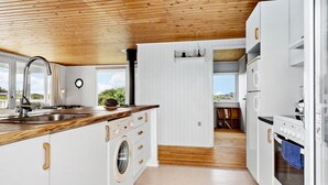 Een compacte keuken met een houten plafond, witte kasten, een ingebouwde wasmachine en uitzicht door een open deur naar een balkon.