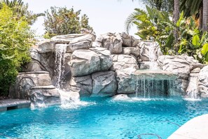 A pool with a waterfall feature surrounded by rocks and tropical plants. - Spacious SoCal Retreat +Pool/Spa Backyard Paradise (Ontario)