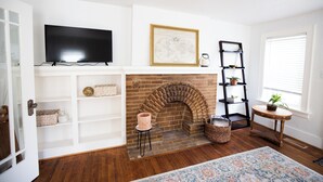 A living room with a brick fireplace, a mounted flat-screen TV, a world map artwork, a bookshelf, and a small round table with a plant. - Charming Oklahoma City Apartment in Central OKC (Oklahoma City)