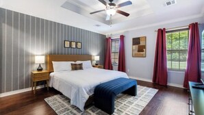 A bedroom with a large bed, a ceiling fan, a nightstand, a window with red curtains, and a view of greenery outside. - Aggie Escape  4BR+Fire Pit + Yard Games Near A&M (College Station)