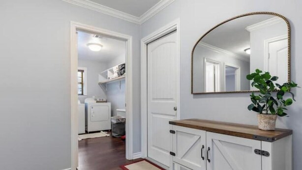 A hallway with a wooden floor, white walls, and a white door. There is a mirror with a gold frame on the wall, and a plant in a woven basket on a wooden shelf. - Aggie Escape  4BR+Fire Pit + Yard Games Near A&M (College Station)